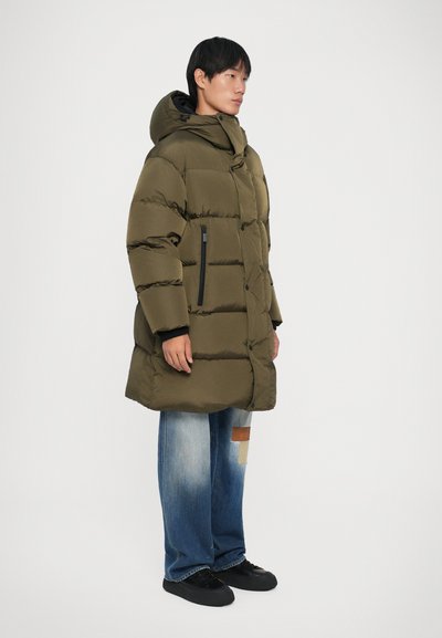 Giacca puffer verde oliva con cappuccio, dotata di zip frontale, tasche con zip nere e polsini elastici, abbinata a jeans blu strappati.