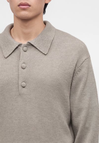 Jeune personne portant un pull en maille beige avec un col et trois boutons couverts de tissu sur un fond uni.