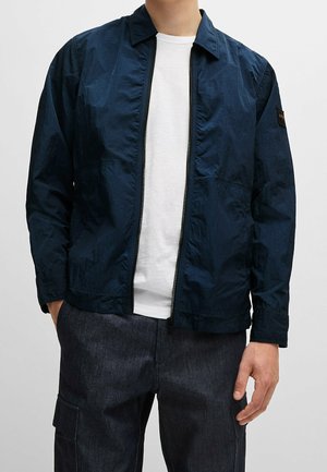 Navyblau leichte Jacke aus einem Ripstop-Material mit einem Frontreißverschluss, Kragen und elastischem Bündchen. Über einem weißen Hemd getragen.