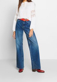 Blouse blanche avec des détails en dentelle sur les manches, associée à des jeans bleus taille haute et à jambes larges, avec une ceinture rouge. Des bottines à talons rouges complètent le look.