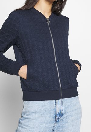 Femme portant une veste bomber zippée bleu marine texturée, les mains dans les poches, et un jean taille haute bleu clair.