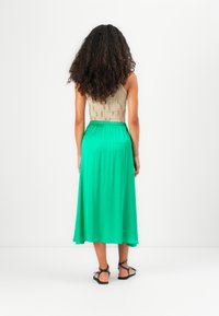 Top bej fără mâneci cu un design texturat, asortat cu o fustă verde lungă și fluidă. Sandale negre cu bretele completează ținuta.