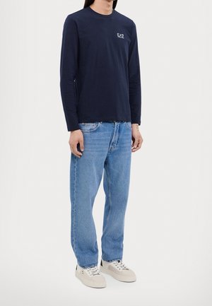 Personne portant un t-shirt à manches longues bleu marine avec logo EA7, un jean bleu clair et des baskets blanches, debout devant un fond uni.