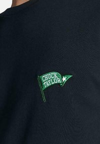 Camiseta de algodón en color azul marino con un pequeño bordado de una bandera verde que lleva el texto "CHUCK TAYLOR" y un acento de estrella, ubicado en la parte izquierda del pecho.
