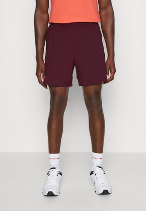 Mężczyzna ubrany w bordowe sportowe szorty, białe skarpetki Nike i białe buty sportowe Nike, stojący na tle jednolitego jasnego tła.