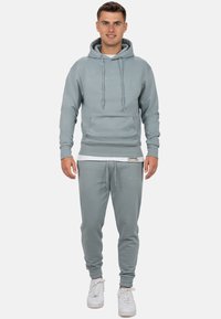Lichtgrijze hoodie met lange koorden, voorzak en bijpassende joggers. Gladde stof, nauwsluitende manchetten en witte sneakers maken de outfit compleet.