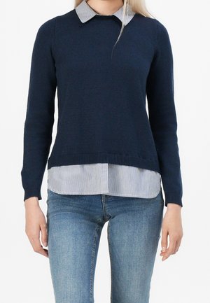 Person wearing a navy blue sweater over a white shirt with thin black stripes and light blue jeans, standing with arms relaxed by their sides.