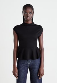 Woman with braided hair wearing a fitted black peplum top and dark blue jeans, standing against a plain white background.