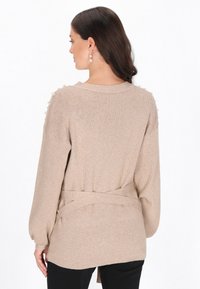 Jasnobrązowy sweter z długim rękawem, z wiązanym paskiem w talii i małymi ozdobnymi koralikami na ramionach. Miękka tekstura.