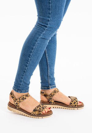 Benedenbenen in blauw skinny jeans met luipaardprint open sandaaltjes met beige zolen, tenen rood gelakt, staand op een witte achtergrond.