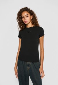 Vrouw met krullend haar, gekleed in een getailleerd zwart T-shirt met een klein wit logo en losse donkere spijkerbroeken, staand tegen een effen achtergrond.
