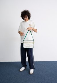White Adidas bag with green strap and gold zipper. Model wears a light t-shirt with flower graphic, dark pants, and white sneakers.