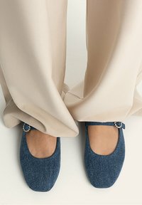 Zapatos planos de ballet de denim azul con punta redondeada y correa, que cuentan con un pequeño detalle decorativo en tono plateado en el costado, usados con pantalones beige.