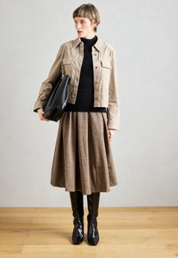 Veste beige raccourcie sur un col roulé noir, associée à une jupe plissée marron et des bottes noires montantes. Sac à main noir à la main.