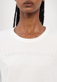 T-shirt in cotone bianco con testo "ARMANI EXCHANGE" in perline argentate. Collo rotondo e texture liscia.
