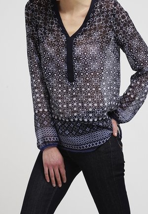 Blusa trasparente blu navy con motivo geometrico bianco, scollatura a V e maniche lunghe con polsini decorati, abbinata a jeans scuri.
