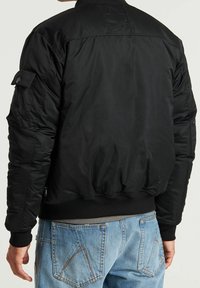 Man in een zwarte bomberjack en lichtblauwe denim jeans, van achteren weergegeven met ontspannen armen langs zijn zij.