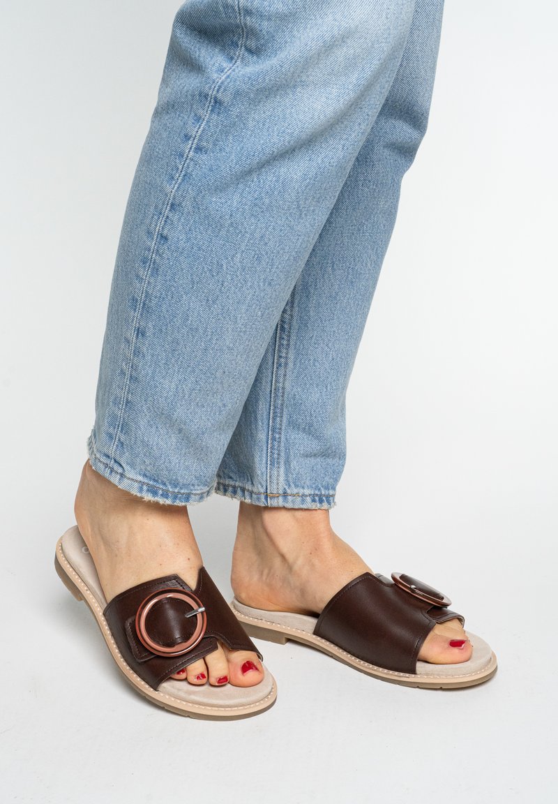 Fødder iført lyseblå jeans og brune åbne slidesandaler med store runde spænder og rødlakerede tånegle på hvid baggrund.