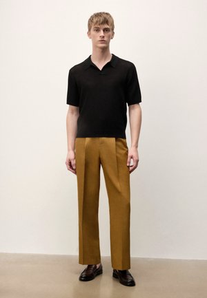 Junger Mann steht, trägt ein schwarzes Strickpolo mit kurzen Ärmeln, senffarbene maßgeschneiderte Hosen und dunkelbraune Loafers, einfarbiger heller Hintergrund.