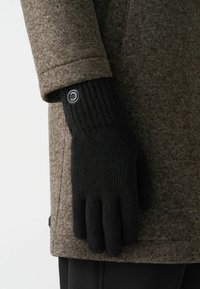 Juodos megztos pirštinės su raštuotais rankogaliais, turinčios apvalų pilką mygtuką, derinamos su ruduo tekstūruotu paltu.