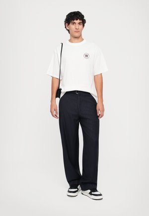 White oversized t-shirt with logo, paired with navy pinstriped pants. Worn with black and white sneakers, and a small black crossbody bag.