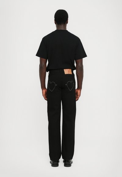 Svarta denimjeans med hjärtformad sömnad på bakfickorna, i kombination med en enkel svart t-shirt. Enfärgad, standard passform, minimala detaljer.