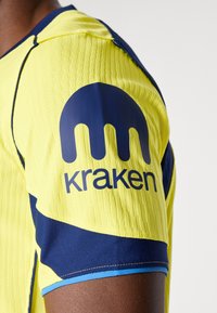 Kortærmet trøje i lys gul med tekstureret stof. Har marineblå accenter og et rundt logo med "kraken" trykt.