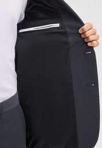 Osoba trzymająca otwartą ciemnoszaro-skalną marynarkę, pokazująca gładką czarną podszewkę i wewnętrzną kieszeń z białym wykończeniem.