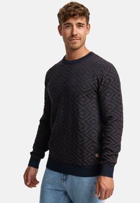 Pull en motif marine avec un design en losanges, col rond et poignets côtelés. Fabriqué en tissu texturé, il présente une petite étiquette sur le côté inférieur.