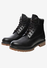 Botas de cuero negro hasta el tobillo con ojales de metal, cordones marrones y una suela de goma con una superficie texturada y detalle del logotipo en el lateral.