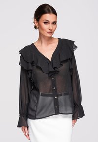Femme portant une blouse noire transparente avec des volants, des boutons dorés et des manches bouffantes, assortie à une jupe blanche, se tenant devant un fond clair.