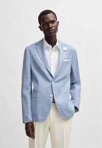 Ljusblå blazer i strukturerat tyg med ett subtilt rutig mönster, med vita knappar, en bröstficka och en vit blomma som accent.