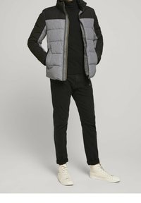 Veste rembourrée en gris et noir avec une fermeture éclair, dotée de poches latérales et d'un col montant. Portée sur un pull noir à col roulé et un pantalon noir.
