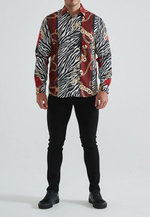 Chemise à manches longues avec des rayures zèbres, des accents rouges et des motifs de chaînes dorées. Un pantalon noir slim-fit et des baskets noires complètent le look.