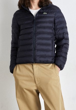 Homme portant une doudoune bleu marine zippée, pantalon beige avec les mains dans les poches, et une chemise blanche en dessous, debout contre un mur uni.