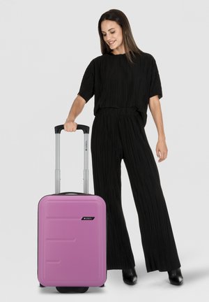 Roze harde koffer met gestructureerd oppervlak, zwarte accenten, uitschuifbare handgreep en spinnerwielen. Model draagt een zwart geplooide outfit.