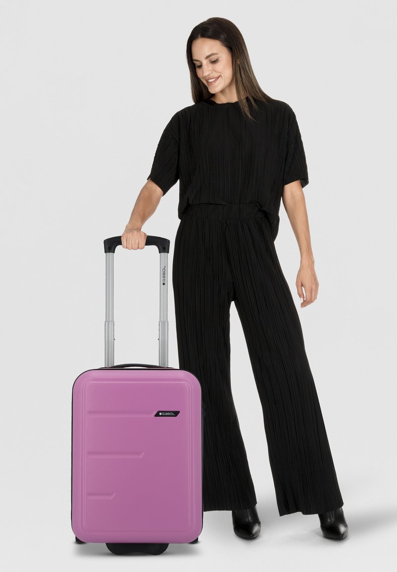 Maleta dura rosa con superficie texturizada, acentos negros, mango retráctil y ruedas giratorias. El modelo lleva puesto un conjunto plisado negro.