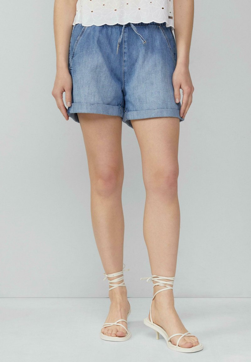 Shorts en denim avec des ourlets roulés, couleur bleu clair, taille élastique avec cordon de serrage. Associés à des sandales blanches à lanières.