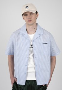 Jeune homme portant une casquette beige, une chemise rayée bleu clair à manches courtes sur un T-shirt blanc avec des logos assortis, et un pantalon foncé, les mains dans les poches.