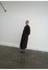 Długi, ciemnobrązowy trench z szerokim kołnierzem, kieszeniami z przodu i luźnym krojem. Noszony na szare rajstopy i czarne obcasy na neutralnym tle.