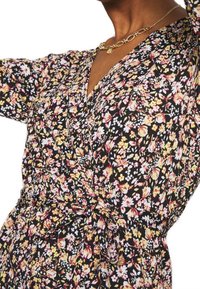 Czarna sukienka w kwiaty z żółtymi, różowymi i białymi kwiatami, z marszczonym pasem i dekoltem w kształcie litery V. Miękka faktura materiału i długie rękawy.