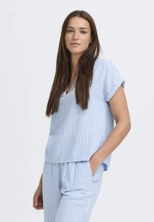 Fiatal nő hosszú barna hajjal, világoskék-fehér csíkos rövid ujjú felsőt és hozzáillő nadrágot visel, egyik kezét a zsebében tartva áll.