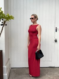 Robe maxi rouge ajustée avec un décolleté arrondi et une découpe sur le côté. Associée à des lunettes de soleil noires et un petit sac à main. Tissu lisse avec une texture minimale.