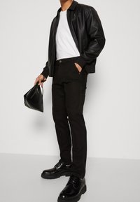 Chaqueta de bomber de cuero negra sobre una camiseta blanca, combinada con pantalones negros. Zapatos negros brillantes, sosteniendo un bolso de mano negro.