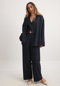 Completo a righe navy con pantaloni a gamba larga e blazer lungo. Tessuto morbido, chiusura con bottone, top corto sotto, abbinato a tacchi beige.