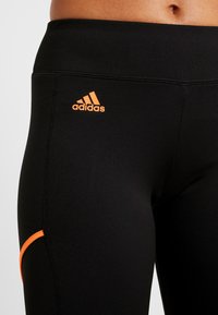 Närbild av svarta Adidas leggings med orange logotyp och orange kant vid låret, som visar midjan och övre benområdet på en hudfärgad modell.