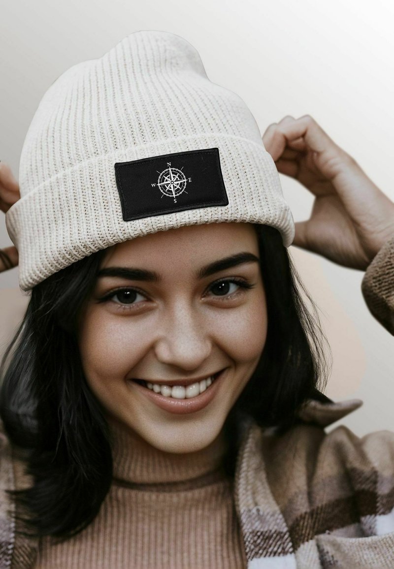 Cream-colored ribbed beanie with a black rectangular patch featuring a white compass design. Soft texture and snug fit.