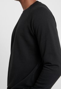 Sudadera negra con cuello redondo, mangas largas y puños de canalé. La tela tiene una textura suave con detalles de costura mínimos.