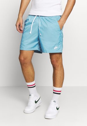 Mężczyzna ubrany w jasnoniebieskie spodenki Nike z białym sznurkiem, białe buty sportowe Nike oraz białe skarpetki z czerwono-czarnymi paskami.