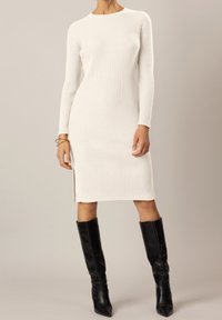 Robe tricotée blanche côtelée avec des manches longues et une fente latérale, dotée d'un col rond. Associée à des bottes noires à talons pointus jusqu'aux genoux.